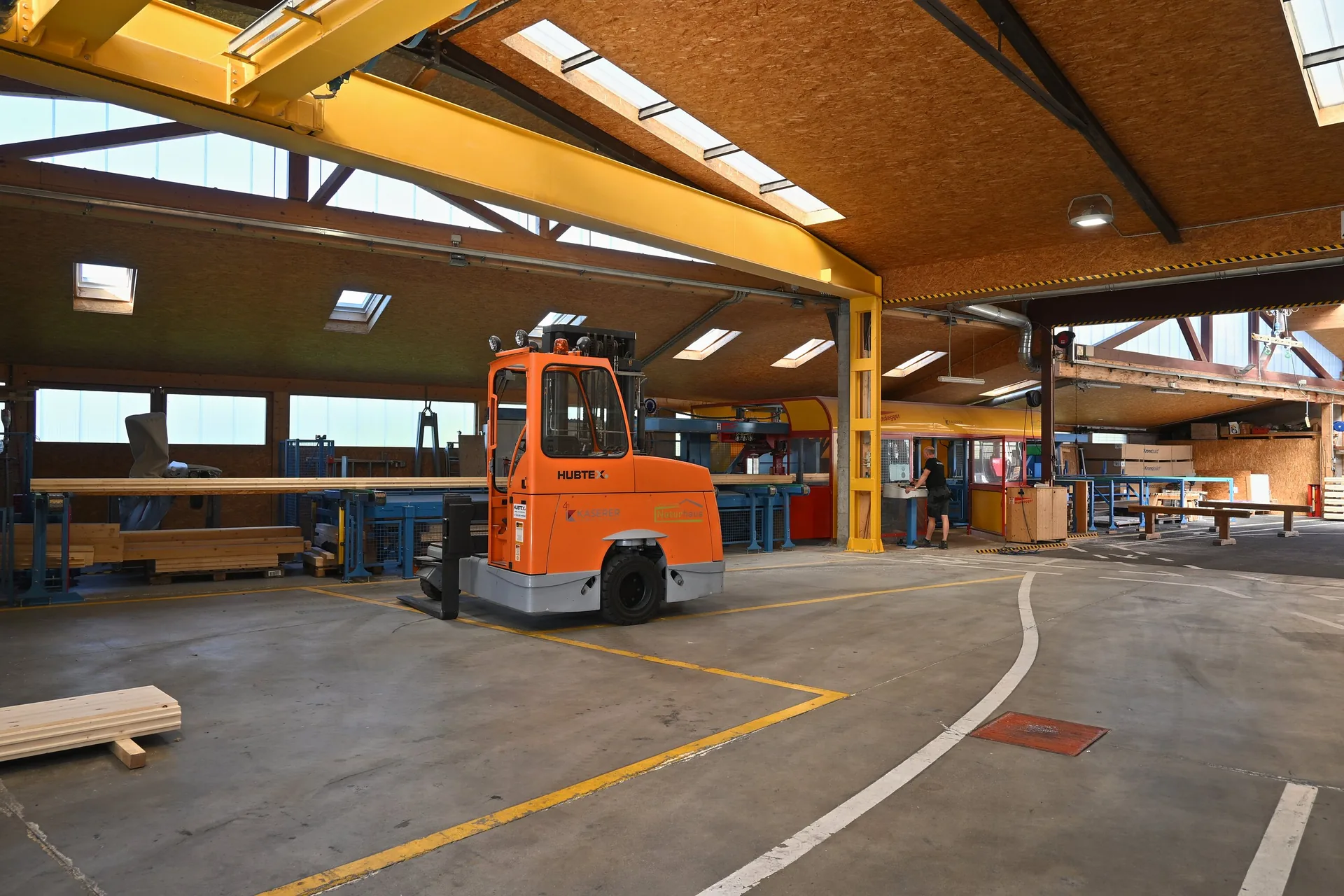 Weite Produktionshalle mit Holzdecke und Oberlichtern, in der Mitte ein orangefarbener Stapler