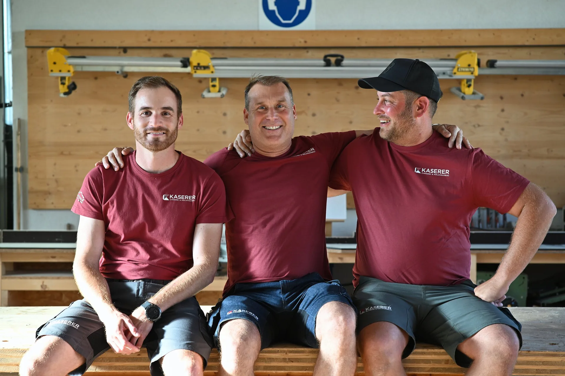 Drei Zimmerer in roten Kaserer-Shirts sitzen lachend Arm in Arm auf einem Holzstapel in der Werkstatt