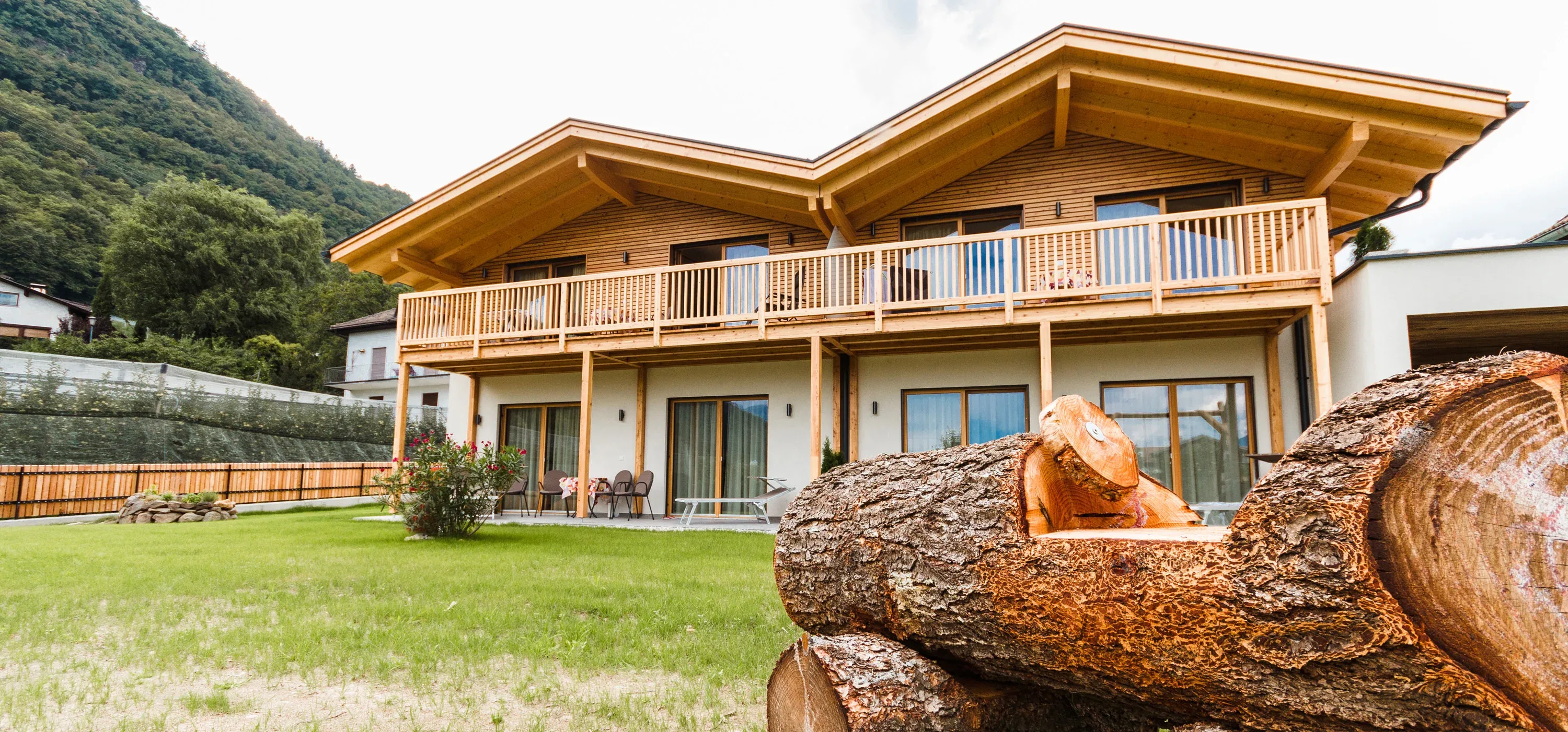 Holzhaus mit durchlaufendem Balkon, im Vordergrund abgelängte Baumstämme