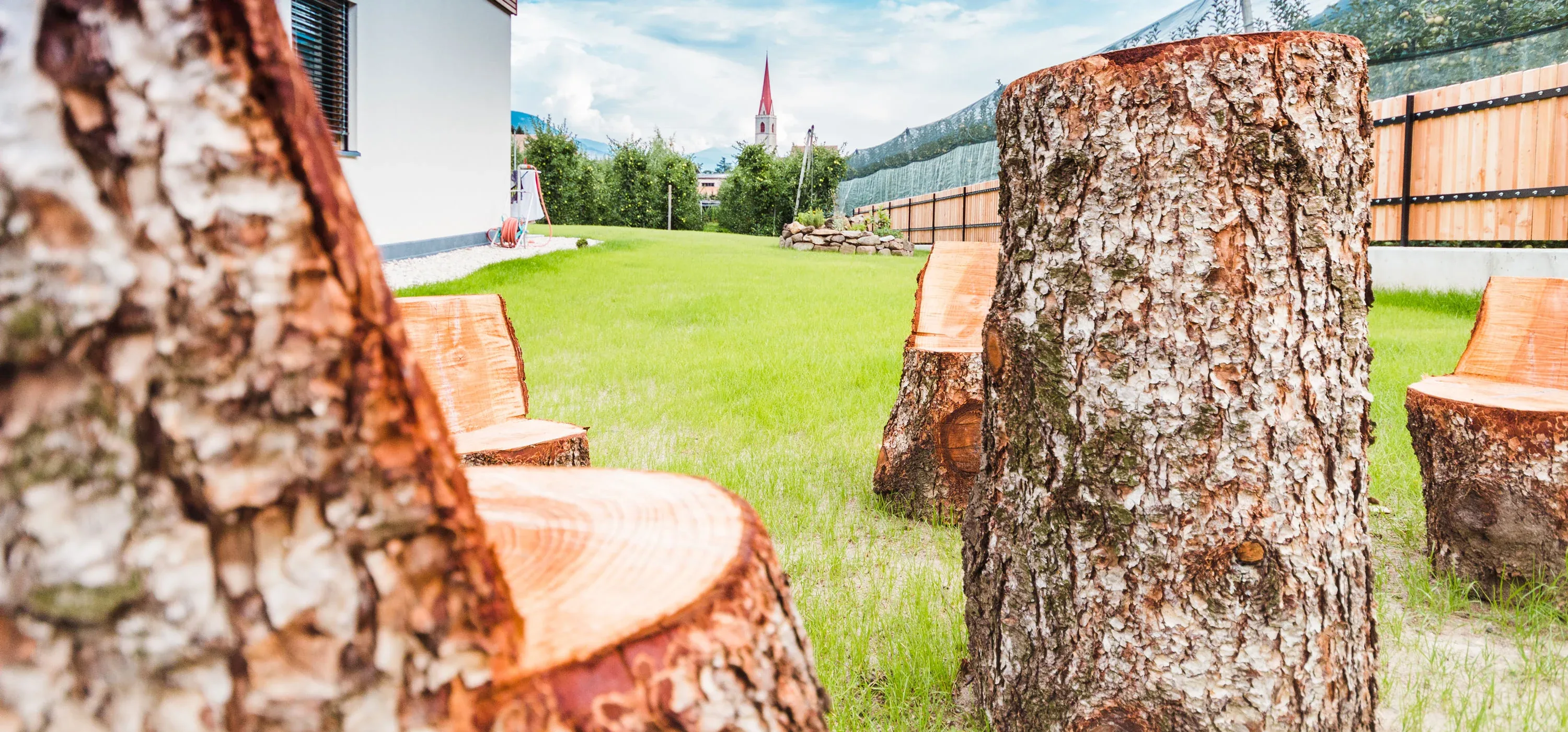 Sessel aus ausgesägten Baumstämmen auf einer Wiese, dahinter ein Kirchturm